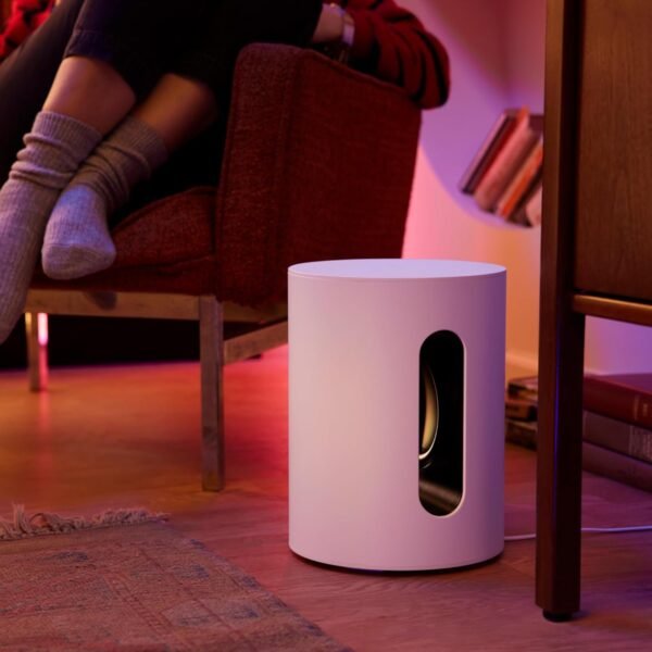 A cylindrical white speaker or subwoofer on the floor next to a red armchair, with a person’s legs resting on the armchair. The room is dimly lit with pink and purple lighting.