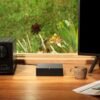 Table with an audio receiver, Sonos device, coffee mug, and monitor stand, with a window view of green plants in the background.