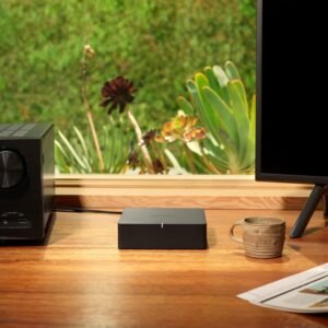 Table with an audio receiver, Sonos device, coffee mug, and monitor stand, with a window view of green plants in the background.