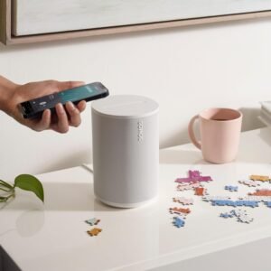 A hand holding a smartphone next to a white Sonos speaker on a table with a puzzle in progress and a pink mug.