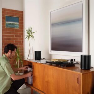 A man is kneeling, selecting records from a wooden cabinet. A turntable and two black speakers are on top of the cabinet. The background includes a brick wall, a plant, and two framed pictures.