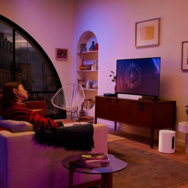 A person sitting on a couch in a dimly lit living room, facing a TV screen with sci-fi graphics. The room features a large arched window, a bookshelf, a white modern chair, and various decor items on a wooden console.