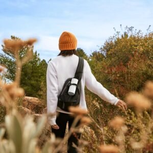 Person wearing a white sweater, orange beanie, and black backpack walking through a natural area with plants and bushes.