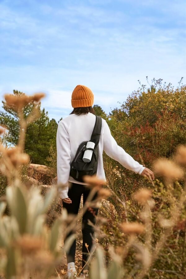 Person wearing a white sweater, orange beanie, and black backpack walking through a natural area with plants and bushes.