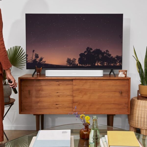 Modern living room with a flat-screen TV displaying a night sky scene, situated on a wooden console with a white soundbar underneath. A person is standing on the left side holding a remote, and various plants and books are placed around the room.